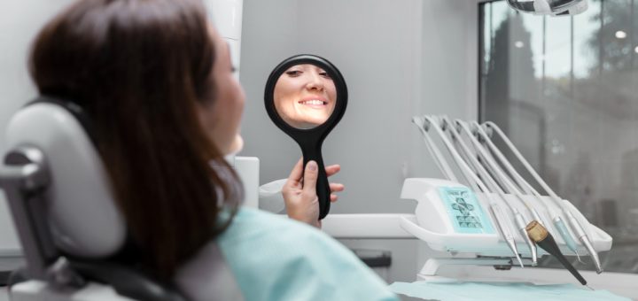 Patient in a dental chair holds a handheld mirror and smiles at her reflection during a checkup, with dental tools on a tray nearby.