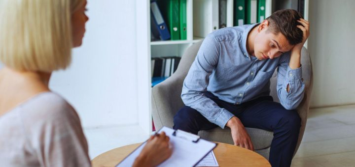 Young man in a blue shirt sits with head in hand in a therapy session while a clinician takes notes on a clipboard.