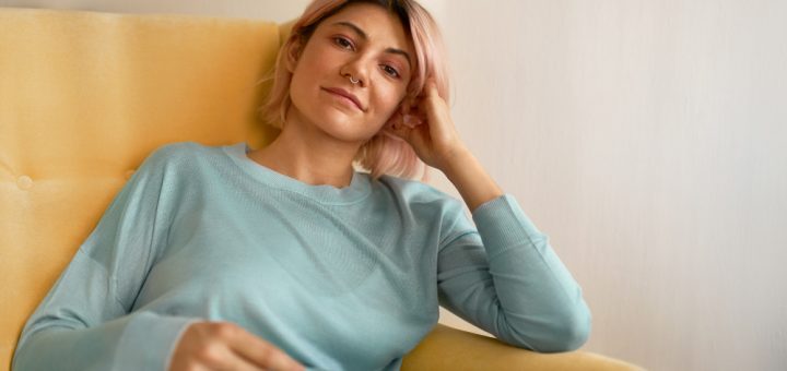 Calm, casual portrait of a person lounging on a mustard-yellow chair in a light blue sweater.