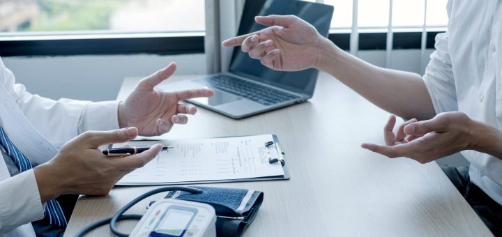Two professionals review medical forms on a desk, with a blood pressure monitor and laptop visible.