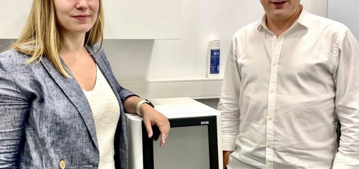 Two professionals pose beside a white medical analyzer with a touchscreen in a clean lab setting.