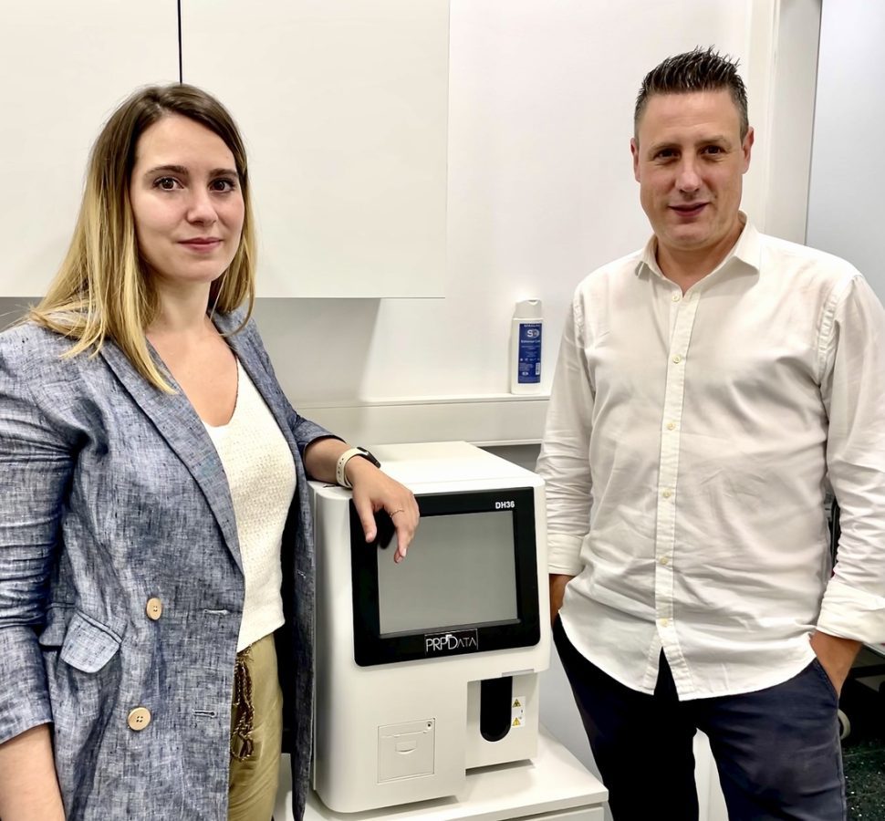 BSD Pain Impact- IP | SoloSalud.net Two professionals pose beside a white medical analyzer with a touchscreen in a clean lab setting.