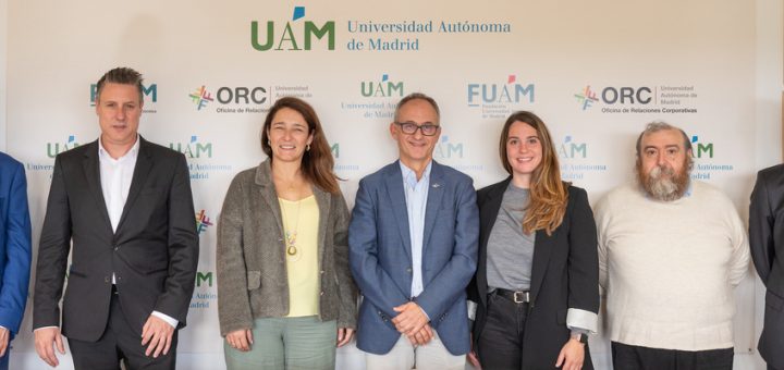 Eight professionals in business attire pose for a group photo in front of a UAM backdrop with logos of Universidad Autónoma de Madrid.