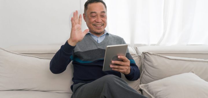 Smiling man sits on a light sofa, holding a tablet and waving hello at the screen.
