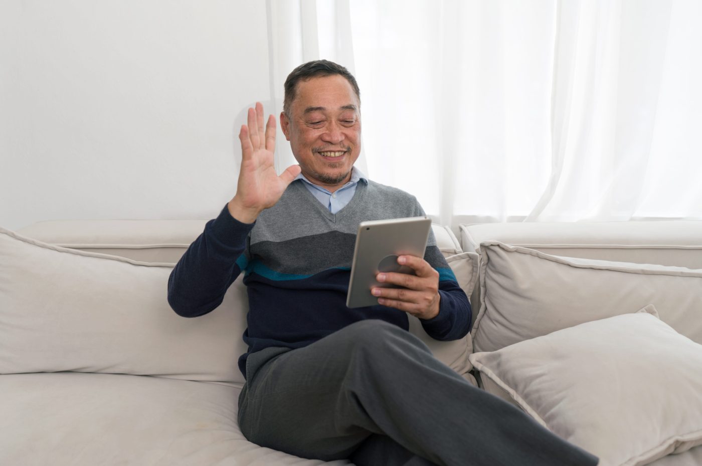 Smiling man sits on a light sofa, holding a tablet and waving hello at the screen.