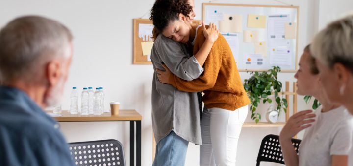 Two coworkers share a comforting hug in a bright meeting room as others sit nearby, suggesting support or consolation in a group setting.