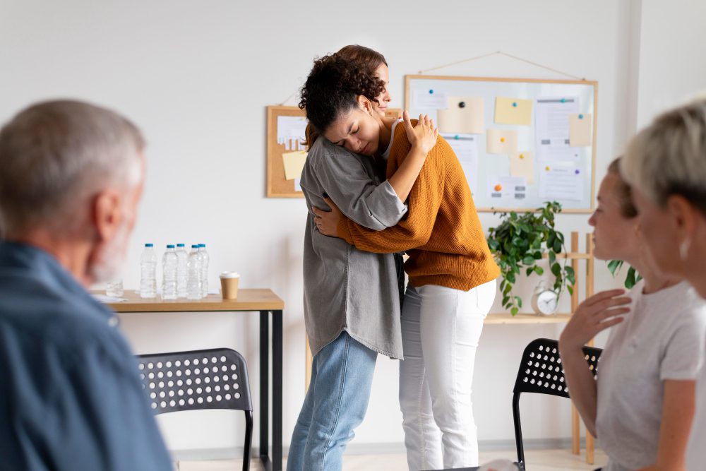 776376 - Mensalud | SoloSalud.net Two coworkers share a comforting hug in a bright meeting room as others sit nearby, suggesting support or consolation in a group setting.