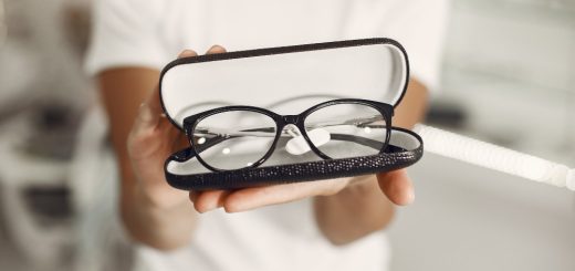 Open glasses case with black-rimmed eyeglasses held toward the camera by a person in a white shirt.