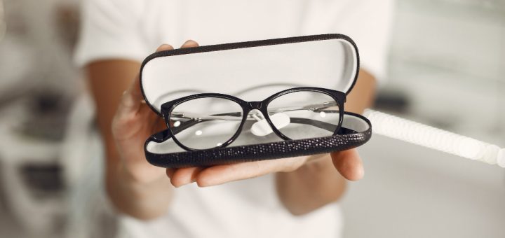 Open glasses case with black-rimmed eyeglasses held toward the camera by a person in a white shirt.