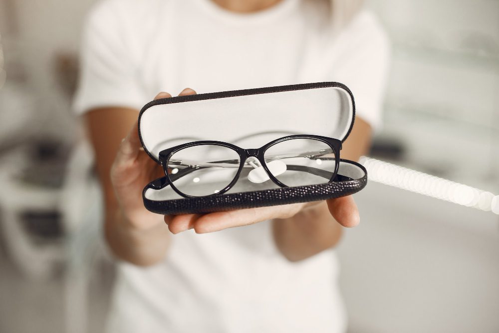 776630 - All Visión | SoloSalud.net Open glasses case with black-rimmed eyeglasses held toward the camera by a person in a white shirt.
