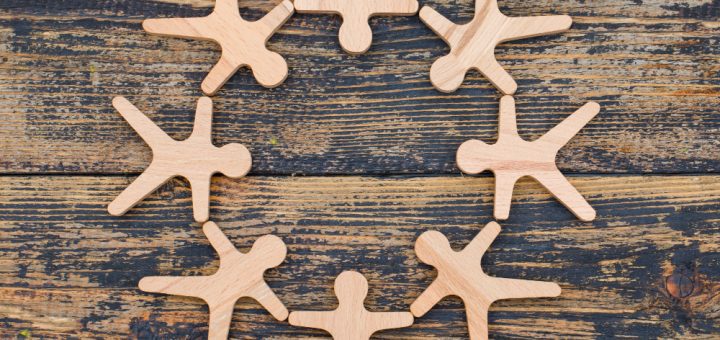Eight wooden figure cutouts form a circle on a dark, weathered wooden surface, like people holding hands around a circle.