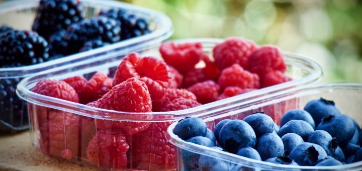 Assorted berries in clear containers: red raspberries, blueberries, and dark blackberries outdoors with a blurred green background.