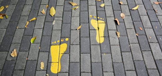 Two yellow footprints painted on a gray brick sidewalk, with fallen leaves around them.