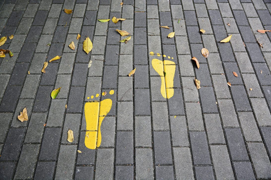 Two yellow footprints painted on a gray brick sidewalk, with fallen leaves around them.