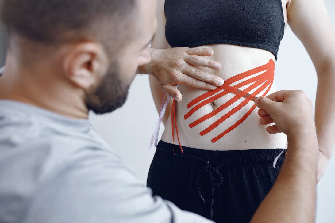 Doctor glues a tepee to an athlete at the hospital | SoloSalud.net Therapist applies red kinesiology tape to a woman's lower abdomen for support or therapy.
