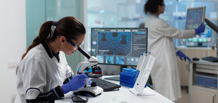 Scientist in a white lab coat uses a microscope at a benchtop, with a computer monitor displaying data nearby.