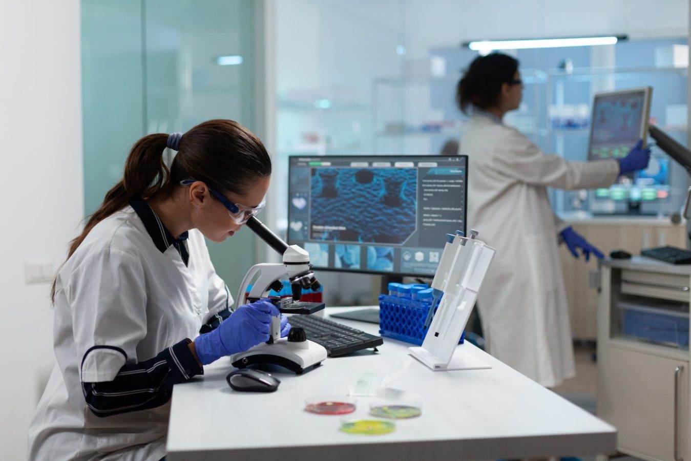 Scientist in a white lab coat uses a microscope at a benchtop, with a computer monitor displaying data nearby.
