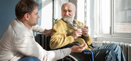 Elegir una residencia para un familiar también implica aprender a acompañarlo - Middle-aged man chats with an elderly man in a wheelchair, both holding a glass in a sunlit room by a large window.