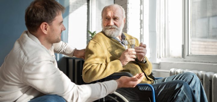 Elegir una residencia para un familiar también implica aprender a acompañarlo - Middle-aged man chats with an elderly man in a wheelchair, both holding a glass in a sunlit room by a large window.