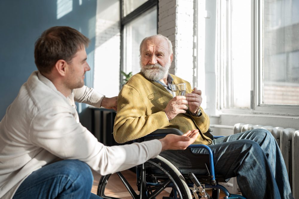 Elegir una residencia para un familiar también implica aprender a acompañarlo - Middle-aged man chats with an elderly man in a wheelchair, both holding a glass in a sunlit room by a large window.