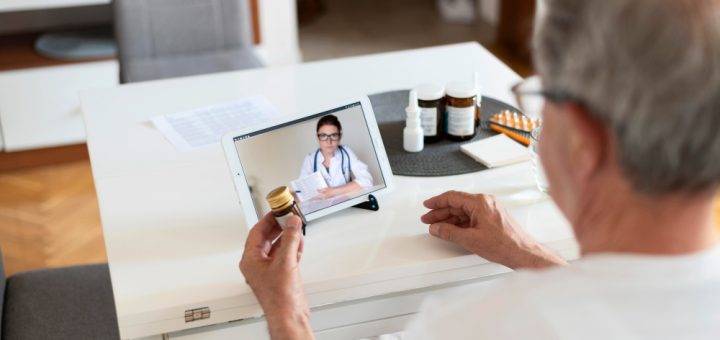 Older man video-calling a doctor on a tablet while holding a medicine bottle at home, with pill bottles on the table nearby