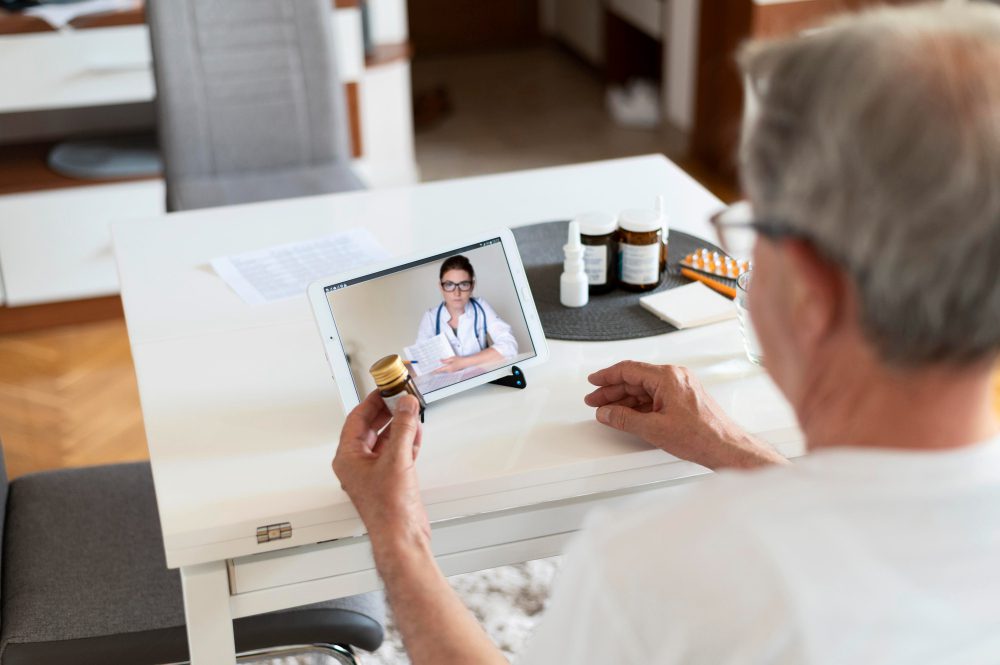 780627 - Home Sweet Home Salud | SoloSalud.net Older man video-calling a doctor on a tablet while holding a medicine bottle at home, with pill bottles on the table nearby
