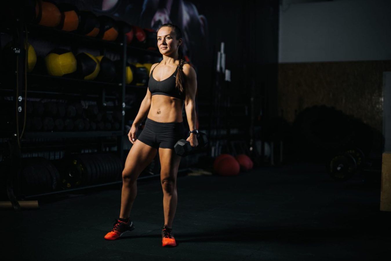 780816 isobiv fold | SoloSalud.net Fit woman in black sports bra and shorts stands in a dim gym, holding a dumbbell in one hand with a focused expression.