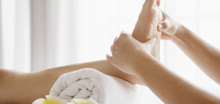 Foot massage in a spa: a therapist massages a bare foot over a rolled towel, with plumeria flowers nearby.