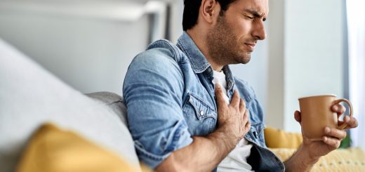 Man sitting on a couch clutching his chest with one hand while holding a mug in the other, looking uncomfortable or in pain.