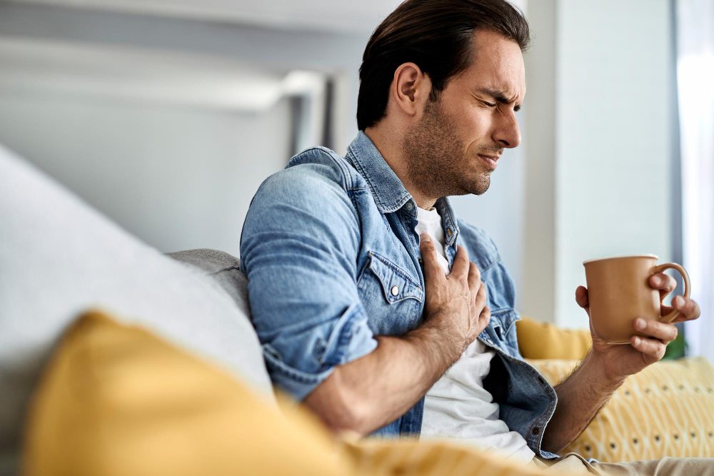 782545 - Clínica Cardiovascular Dr Valderrama | SoloSalud.net Man sitting on a couch clutching his chest with one hand while holding a mug in the other, looking uncomfortable or in pain.