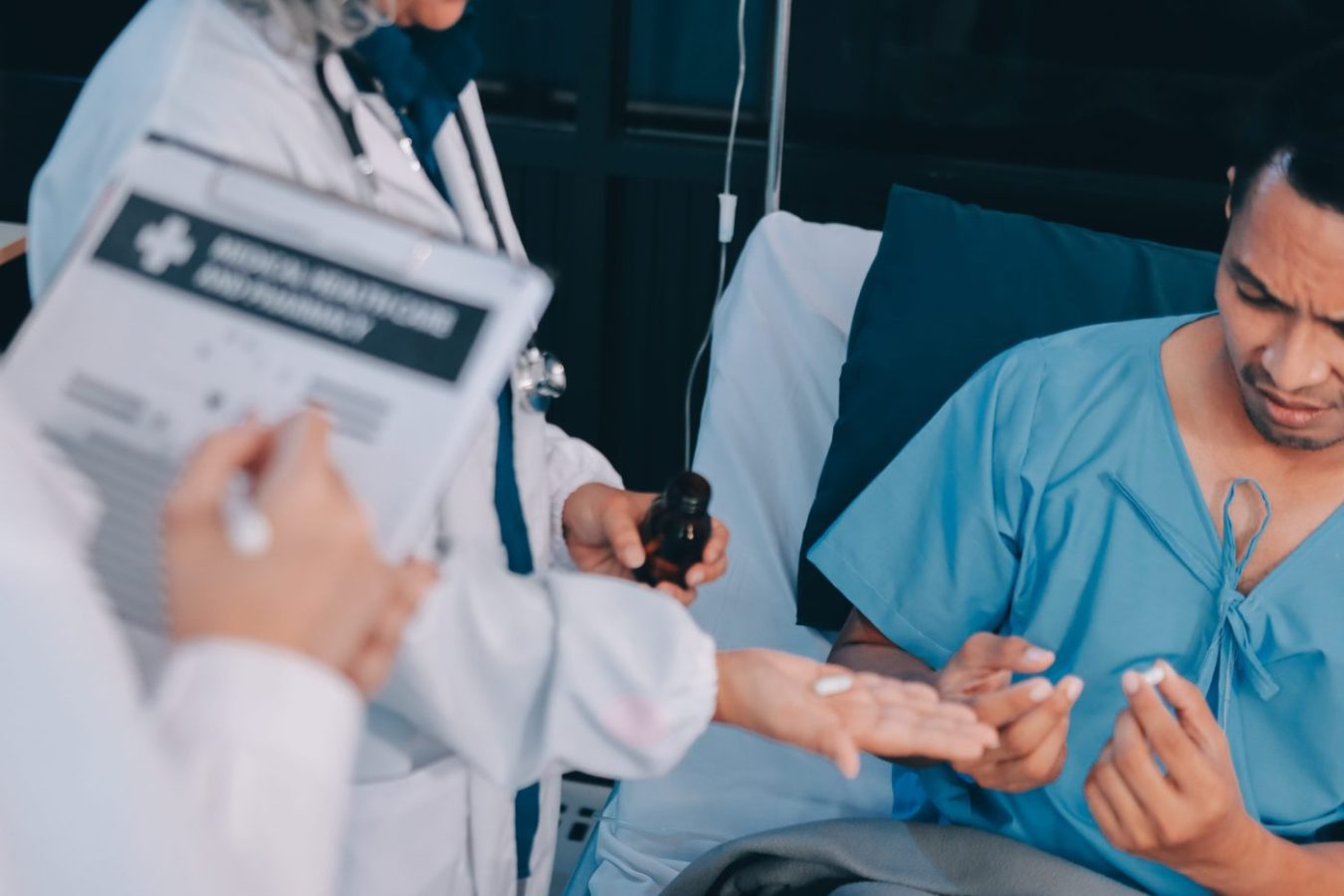 Doctor in white coat with clipboard and bottle explains medication to a male patient in a hospital bed, who holds pills in his hand.
