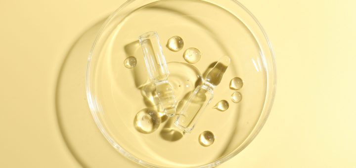 Clear glass ampoules in a shallow dish with yellow background, likely cosmetic serum bottles.