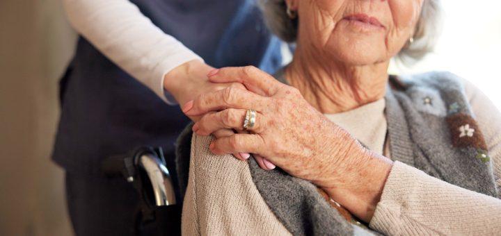Close-up of an elderly woman's clasped hands with a caregiver's hand resting on top, signaling support.