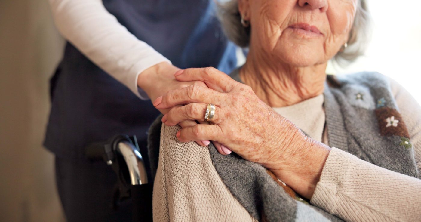 Close-up of an elderly woman's clasped hands with a caregiver's hand resting on top, signaling support.