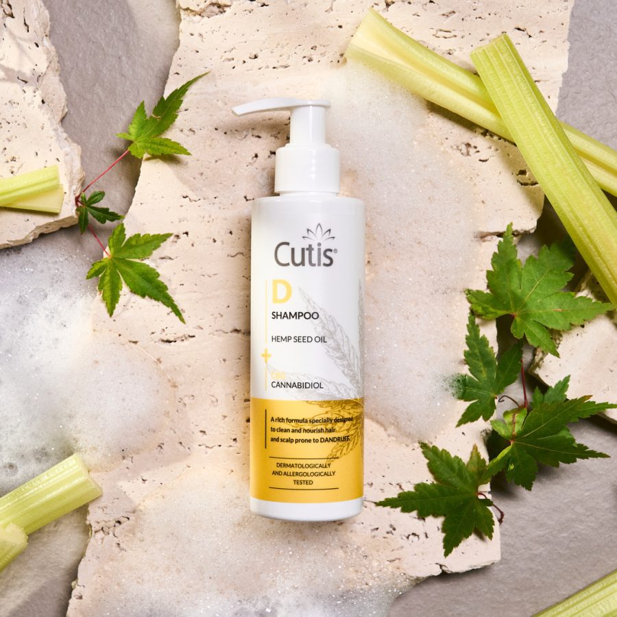 Cutis D Shampoo bottle with hemp seed oil and cannabidiol on a textured surface with green leaves around it (informational product shot)