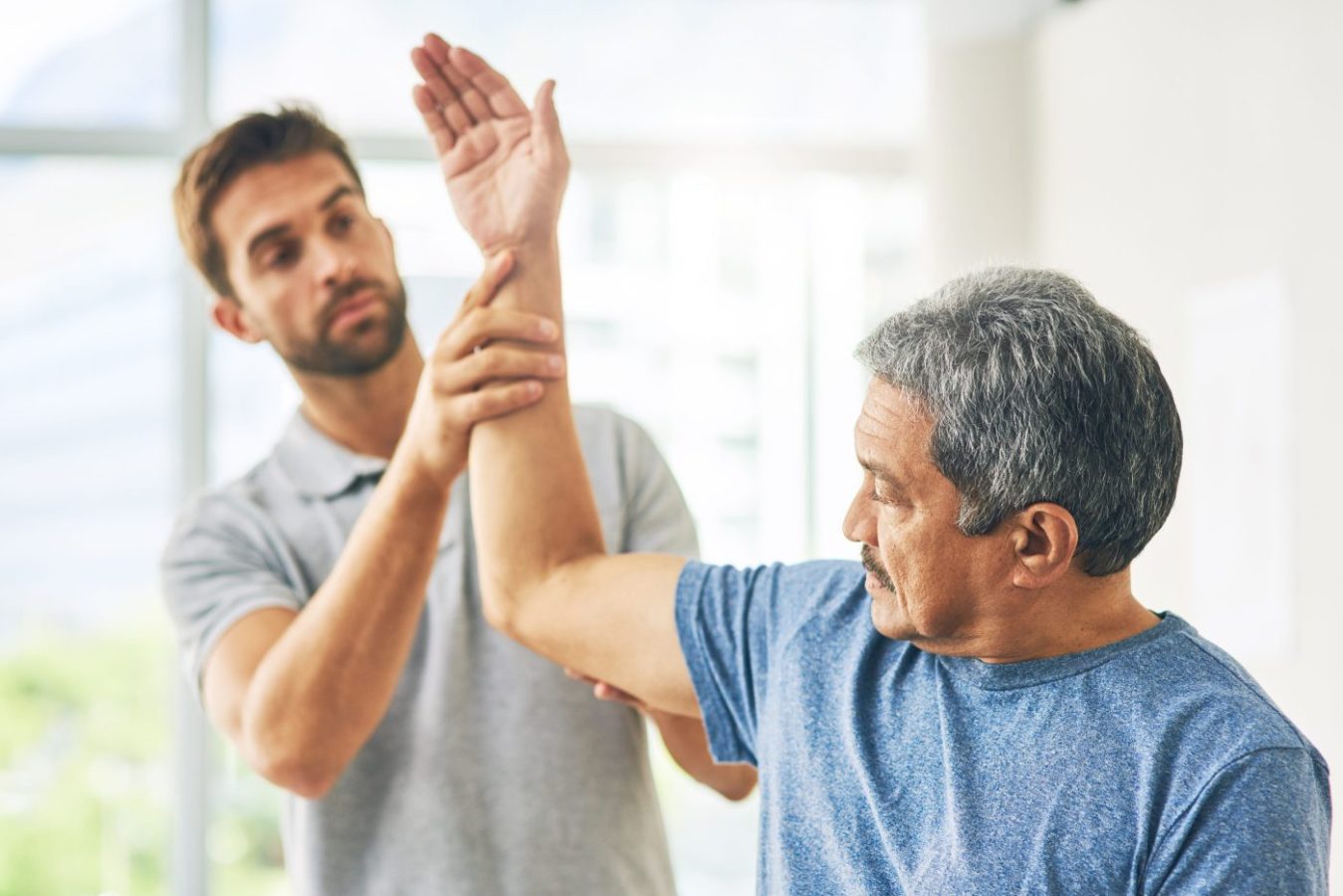 784334 Cefire | SoloSalud.net Therapist guiding a senior man through an elbow stretch in a bright clinic.
