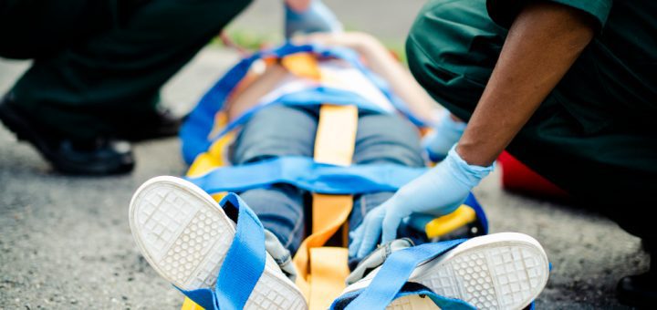 Paramedics secure a patient on a yellow stretcher with blue straps, wearing gloves and kneeling on a paved surface.