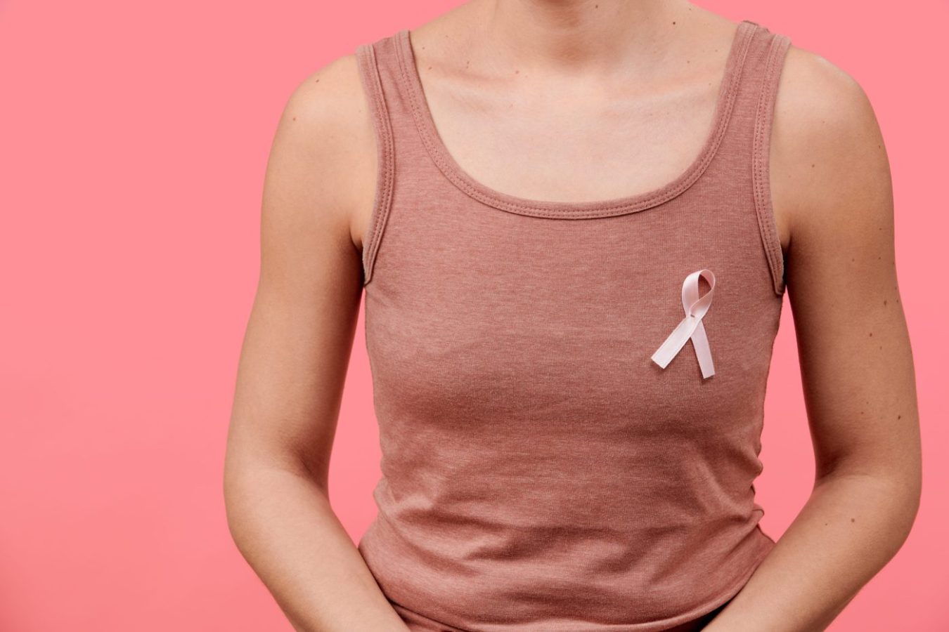 Person wearing a pink tank top with a pink breast cancer awareness ribbon pinned to the chest against a pink background for awareness.